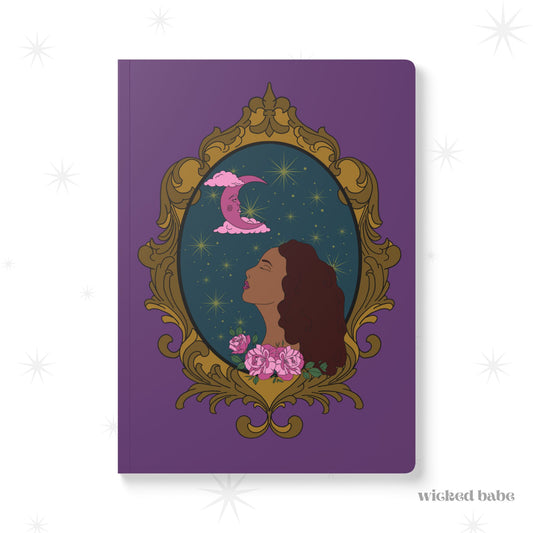 Front cover of purple softcover journal notebook with design of a tan skinned woman with red brown wavy hair with pink flowers and a pink crescent moon above on a teal background surrounded by an ornate gold frame