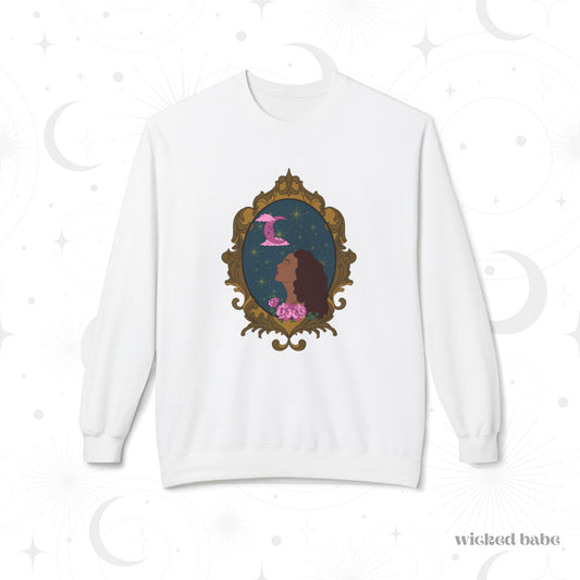 White long sleeve crewneck sweatshirt with design of a tan skinned woman with red brown wavy hair with pink flowers and a pink crescent moon above on a teal background surrounded by an ornate gold frame