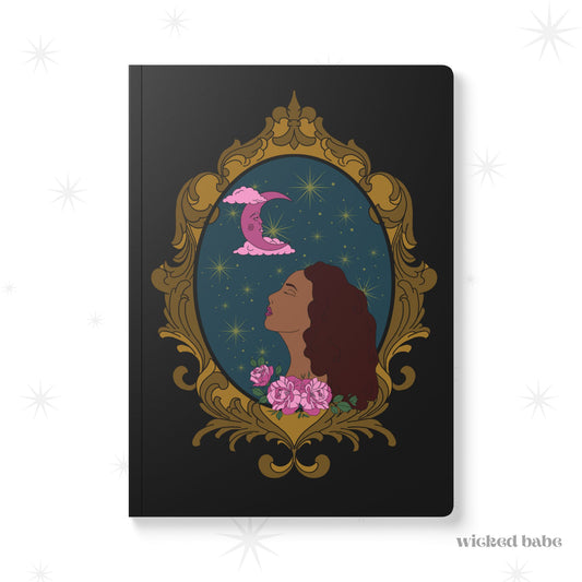 Front cover of black softcover journal notebook with design of a tan skinned woman with red brown wavy hair with pink flowers and a pink crescent moon above on a teal background surrounded by an ornate gold frame