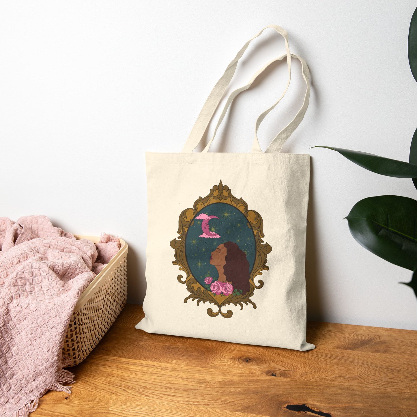 Canvas tote bag with design of a tan skinned woman with red brown wavy hair with pink flowers and a pink crescent moon above on a teal background surrounded by an ornate gold frame leaning against the wall