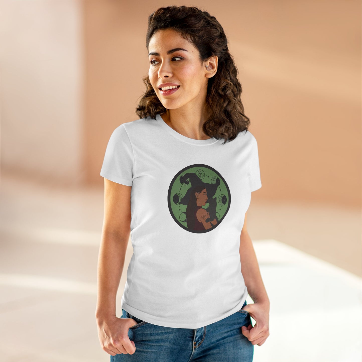 Woman wearing a white short sleeve crew neck t-shirt featuring a design of a woman wearing a witch's hat holding a gray cat with moon phases on a green background