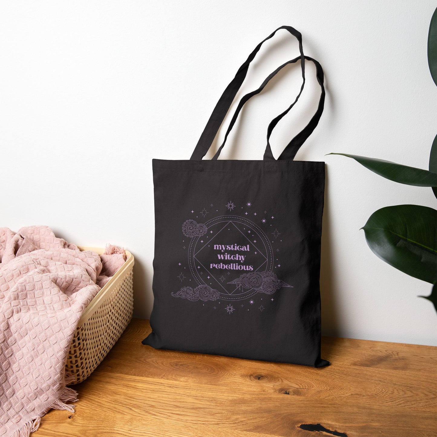 Black tote bag with mystical witchy rebellious text in purple surrounded by clouds and stars leaning against a wall