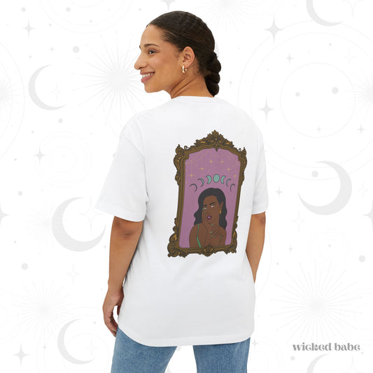 Woman wearing an oversized short sleeve white t-shirt with design of a dark skinned woman with black hair wearing green on a pink background with moon phases above surrounded by ornate gold frame on the back