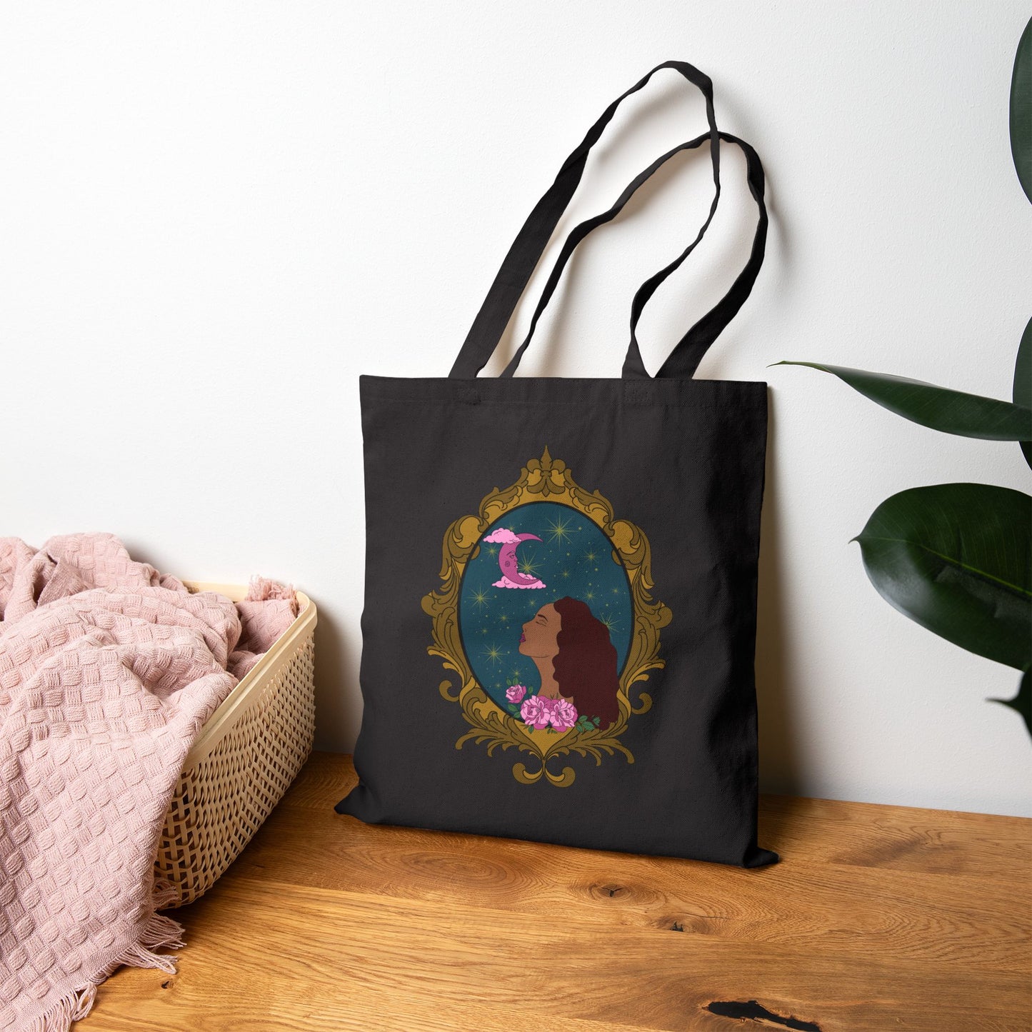 Black tote bag with design of a tan skinned woman with red brown wavy hair with pink flowers and a pink crescent moon above on a teal background surrounded by an ornate gold frame leaning against wall