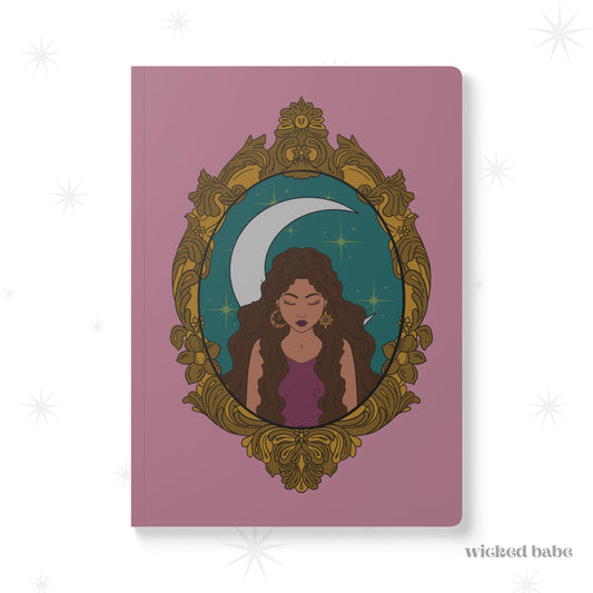 Front cover of pink softcover journal notebook with design of a tan skinned woman with brown wavy hair wearing purple in front of large crescent moon on a teal background surrounded by ornate gold frame