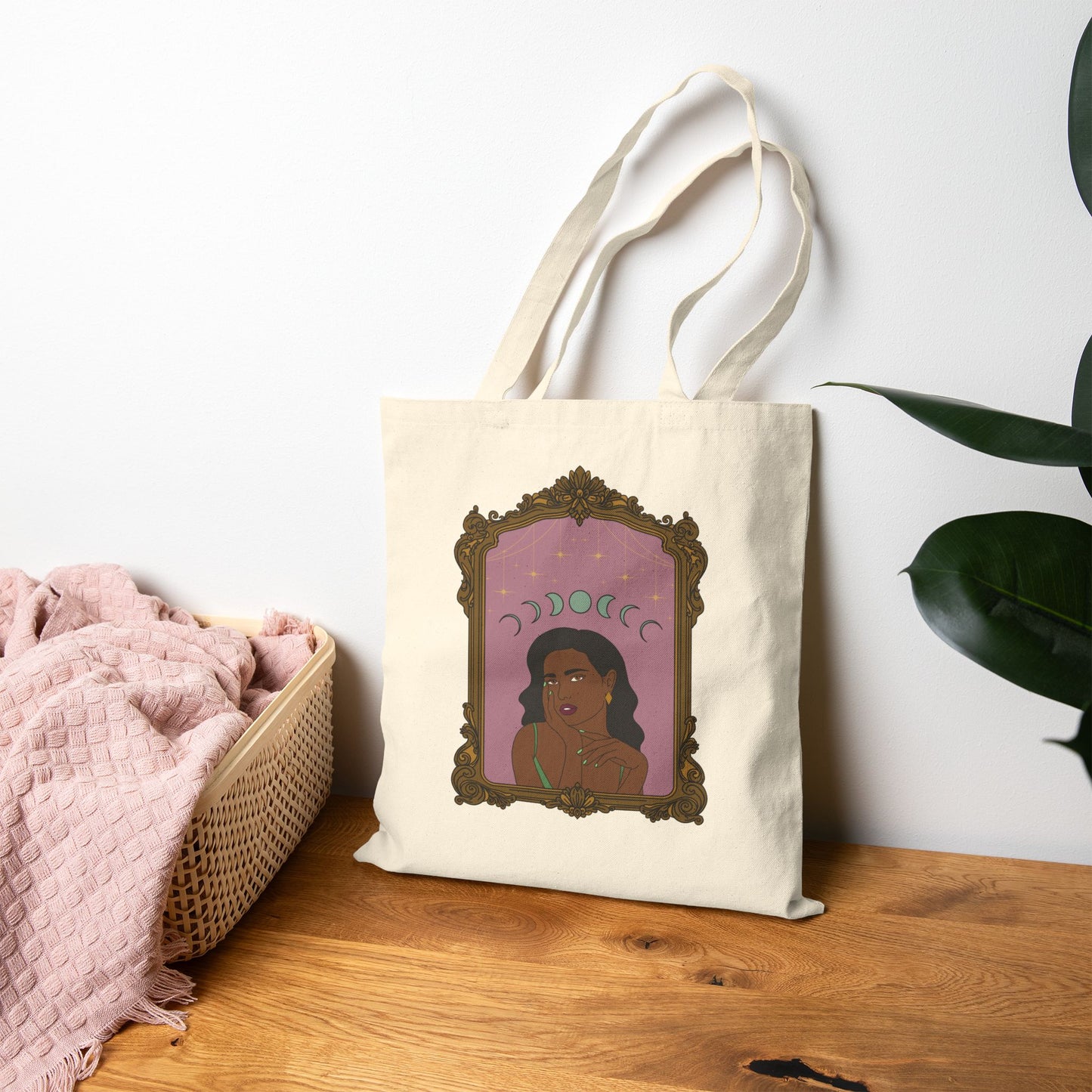 Canvas tote bag with design of a dark skinned woman with black hair wearing green on a pink background with moon phases above surrounded by ornate gold frame leaning against the wall