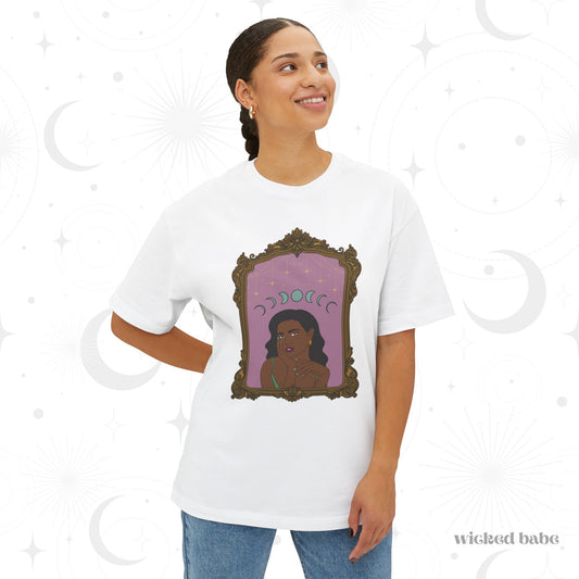 Woman wearing an oversized short sleeve white t-shirt with design of a dark skinned woman with black hair wearing green on a pink background with moon phases above surrounded by ornate gold frame