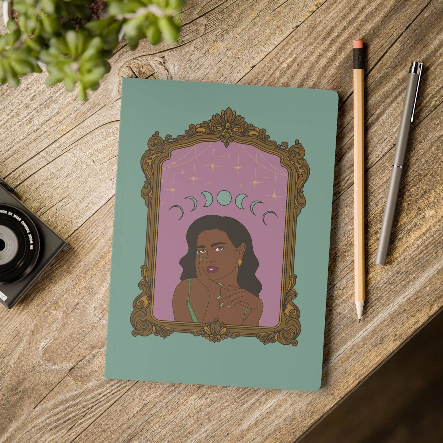 Front cover of green softcover journal notebook with design of a dark skinned woman with black hair wearing green on a pink background with moon phases above surrounded by ornate gold frame on wooden table with pen and pencil