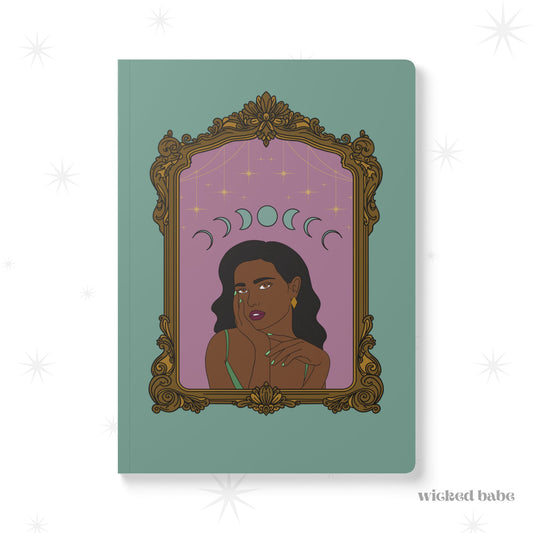 Front cover of green softcover journal notebook with design of a dark skinned woman with black hair wearing green on a pink background with moon phases above surrounded by ornate gold frame