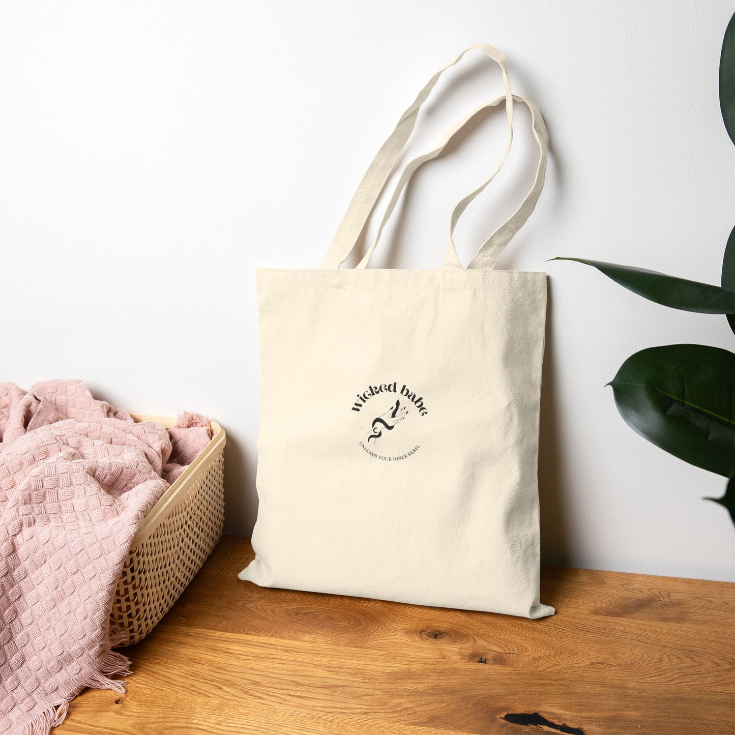 The back of a canvas tote bag with black wicked babe hand with serpent logo in the center leaning against the wall