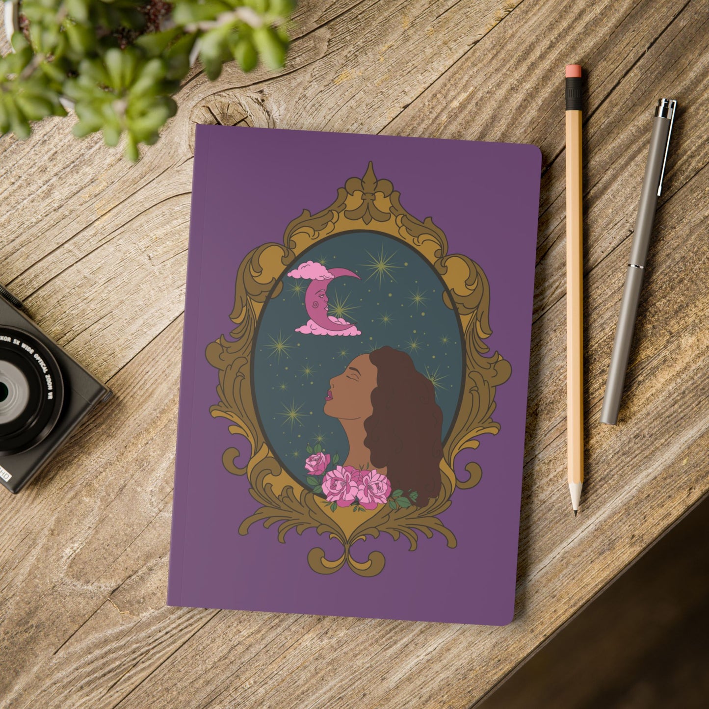 Front cover of purple softcover journal notebook with design of a tan skinned woman with red brown wavy hair with pink flowers and a pink crescent moon above on a teal background surrounded by an ornate gold frame on wooden table with pen and pencil