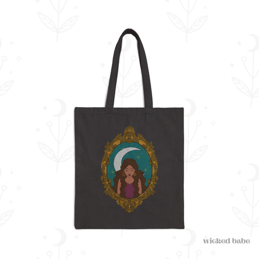 Black tote bag with design of a tan skinned woman with brown wavy hair wearing purple in front of large crescent moon on a teal background surrounded by ornate gold frame