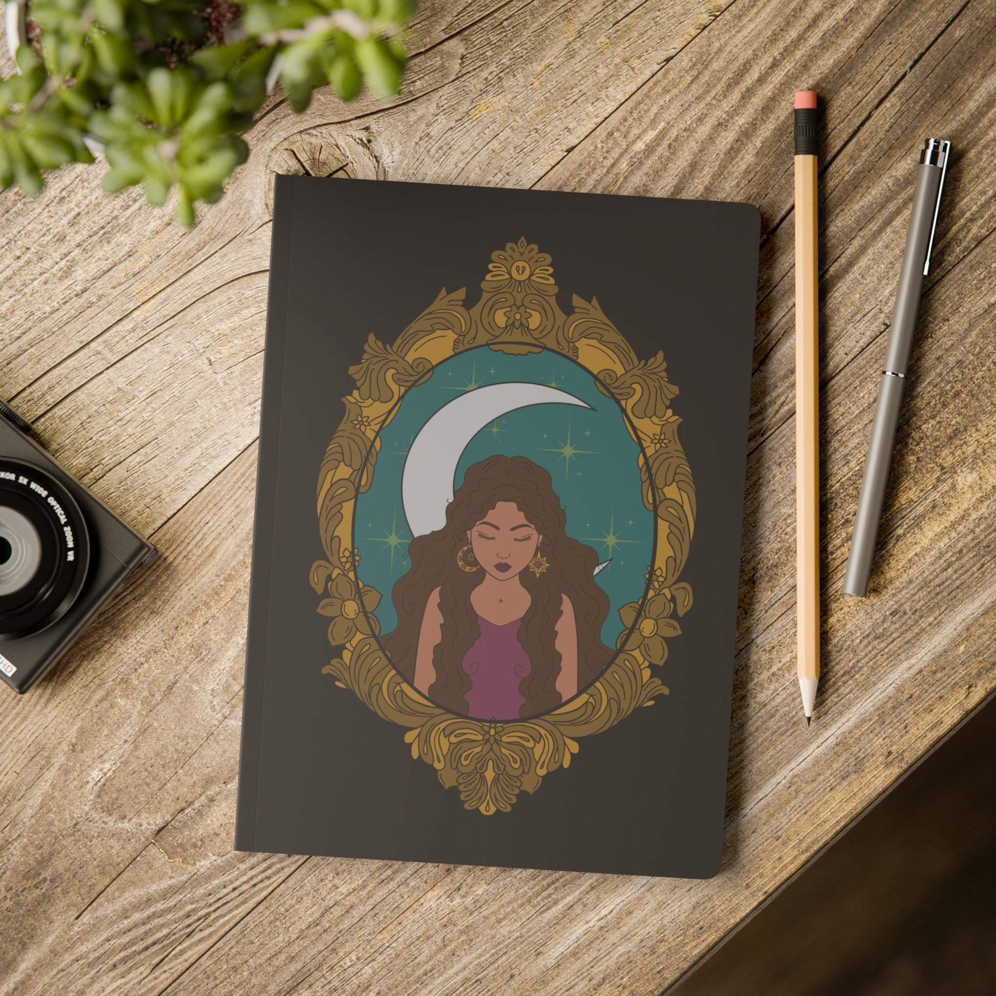Front cover of black softcover journal notebook with design of a tan skinned woman with brown wavy hair wearing purple in front of large crescent moon on a teal background surrounded by ornate gold frame on wooden table with pen and pencil