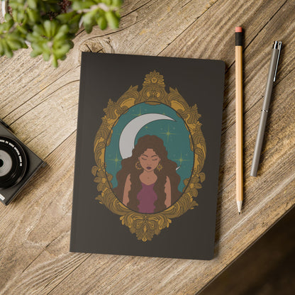Front cover of black softcover journal notebook with design of a tan skinned woman with brown wavy hair wearing purple in front of large crescent moon on a teal background surrounded by ornate gold frame on wooden table with pen and pencil
