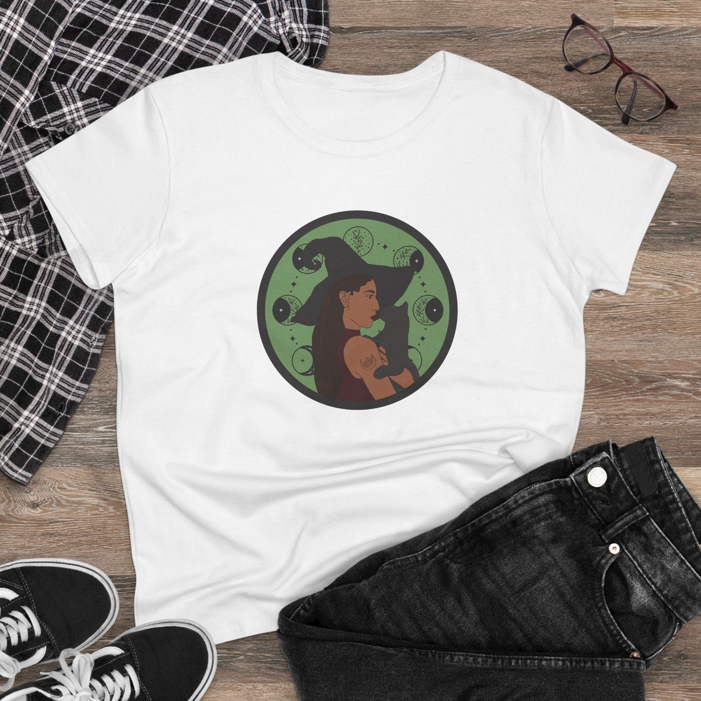 White short sleeve crew neck t-shirt featuring a design of a woman wearing a witch's hat holding a gray cat with moon phases on a green background, displayed as a flatlay with jeans and sneakers