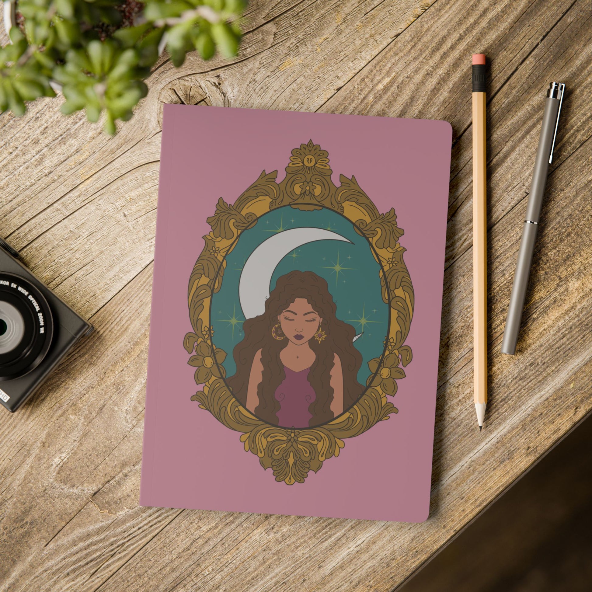 Front cover of pink softcover journal notebook with design of a tan skinned woman with brown wavy hair wearing purple in front of large crescent moon on a teal background surrounded by ornate gold frame on wooden table with pen and pencil