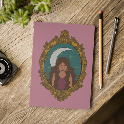 Front cover of pink softcover journal notebook with design of a tan skinned woman with brown wavy hair wearing purple in front of large crescent moon on a teal background surrounded by ornate gold frame on wooden table with pen and pencil