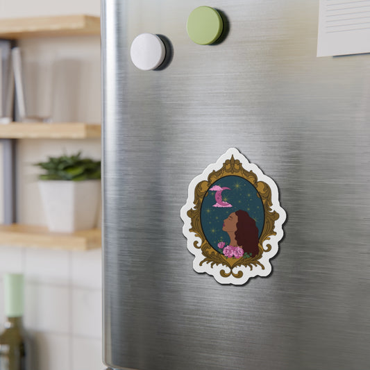 Refrigerator magnet with design of a tan skinned woman with red brown wavy hair with pink flowers and a pink crescent moon above on a teal background surrounded by an ornate gold frame with a white border on a stainless steel fridge