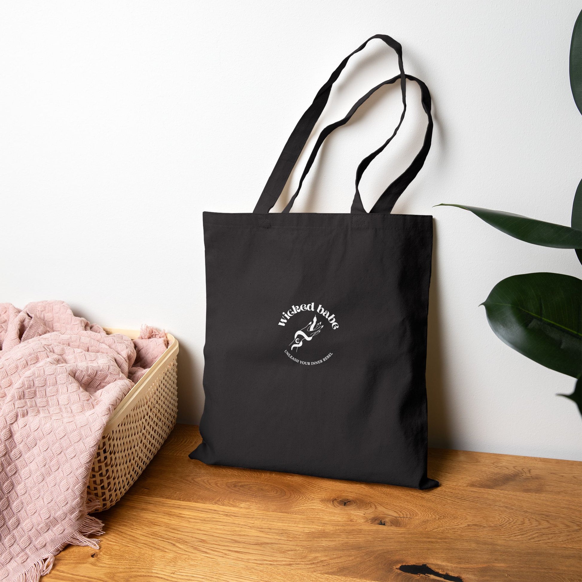 The back of a black tote bag with white wicked babe hand with serpent logo in the center leaning against the wall