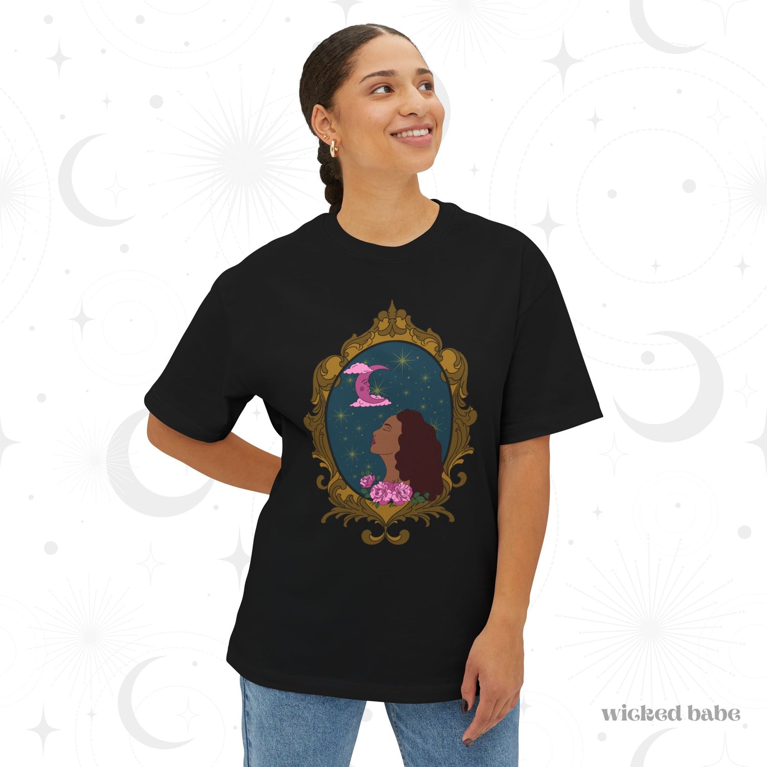 Woman wearing an oversized short sleeve black t-shirt with a design of a tan skinned woman with red brown wavy hair with pink flowers and a pink crescent moon above on a teal background surrounded by an ornate gold frame