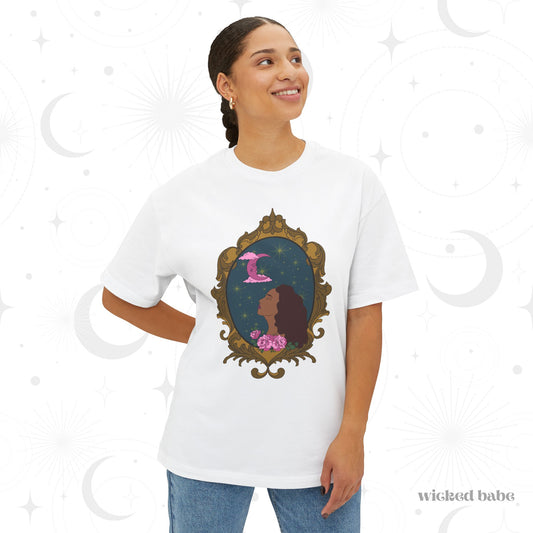 Woman wearing an oversized short sleeve white t-shirt with a design of a tan skinned woman with red brown wavy hair with pink flowers and a pink crescent moon above on a teal background surrounded by an ornate gold frame