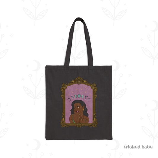 Black tote bag with design of a dark skinned woman with black hair wearing green on a pink background with moon phases above surrounded by ornate gold frame