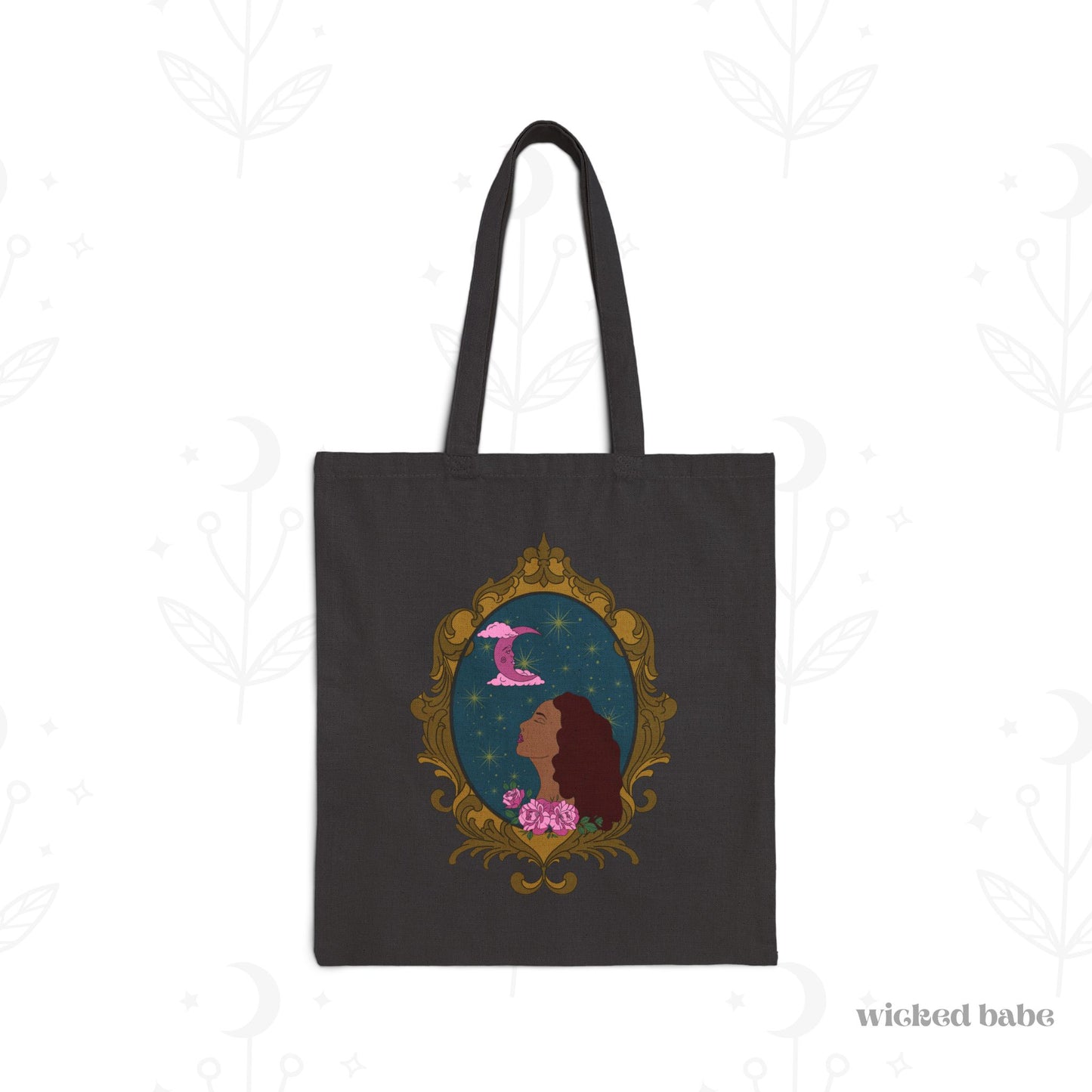 Black tote bag with design of a tan skinned woman with red brown wavy hair with pink flowers and a pink crescent moon above on a teal background surrounded by an ornate gold frame