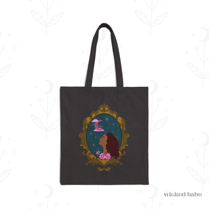 Black tote bag with design of a tan skinned woman with red brown wavy hair with pink flowers and a pink crescent moon above on a teal background surrounded by an ornate gold frame