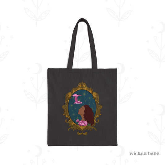 Black tote bag with design of a tan skinned woman with red brown wavy hair with pink flowers and a pink crescent moon above on a teal background surrounded by an ornate gold frame