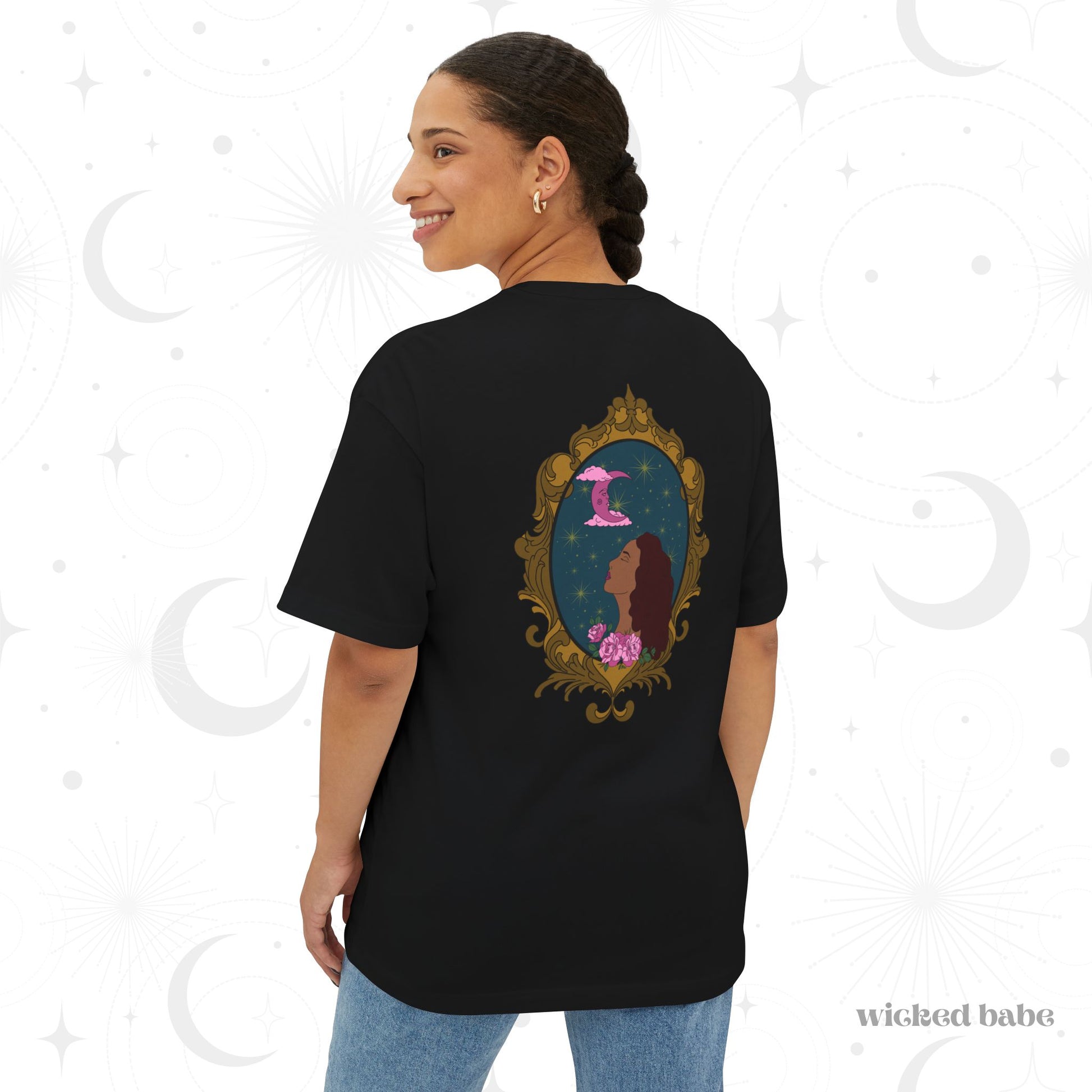 Woman wearing an oversized short sleeve black t-shirt with a design of a tan skinned woman with red brown wavy hair with pink flowers and a pink crescent moon above on a teal background surrounded by an ornate gold frame on the back