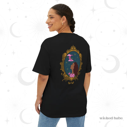 Woman wearing an oversized short sleeve black t-shirt with a design of a tan skinned woman with red brown wavy hair with pink flowers and a pink crescent moon above on a teal background surrounded by an ornate gold frame on the back
