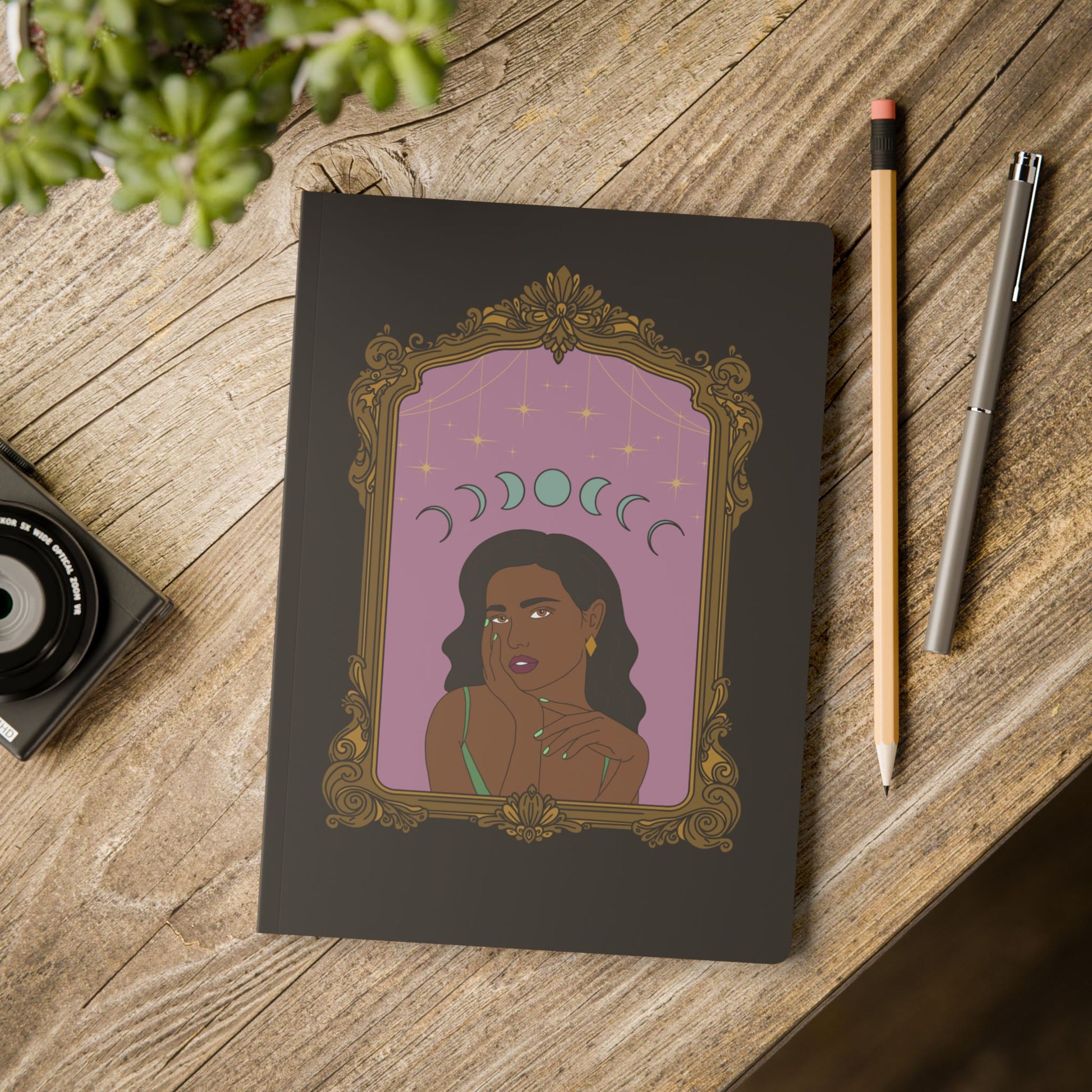 Front cover of black softcover journal notebook with design of a dark skinned woman with black hair wearing green on a pink background with moon phases above surrounded by ornate gold frame on wooden table with pen and pencil