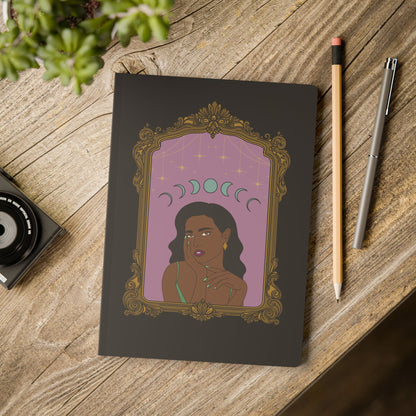 Front cover of black softcover journal notebook with design of a dark skinned woman with black hair wearing green on a pink background with moon phases above surrounded by ornate gold frame on wooden table with pen and pencil