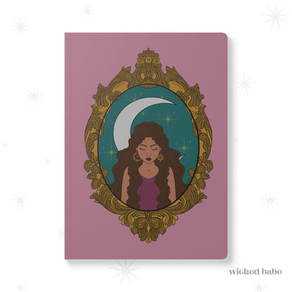 Front cover of pink softcover journal notebook with design of a tan skinned woman with brown wavy hair wearing purple in front of large crescent moon on a teal background surrounded by ornate gold frame