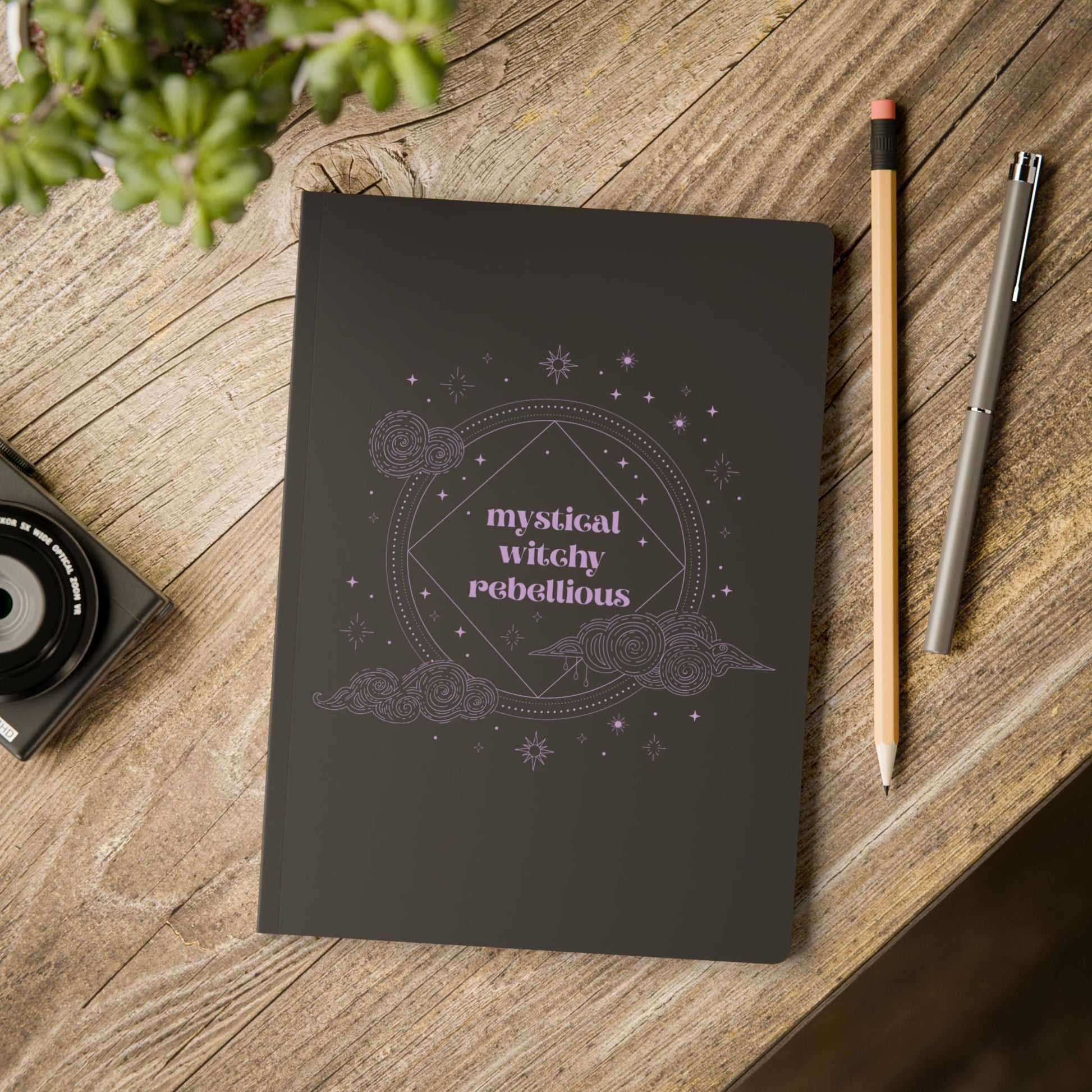 Black softcover journal notebook with mystical witchy rebellious text in purple surrounded by clouds and stars on wooden table with pen and pencil
