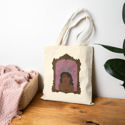 Canvas tote bag with design of a dark skinned woman with black hair wearing green on a pink background with moon phases above surrounded by ornate gold frame leaning against the wall