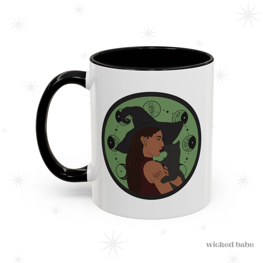 White mug with black accents, handle on left, featuring a design of a woman wearing a witch's hat holding a gray cat with moon phases on a green background