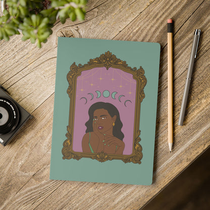 Front cover of green softcover journal notebook with design of a dark skinned woman with black hair wearing green on a pink background with moon phases above surrounded by ornate gold frame on wooden table with pen and pencil