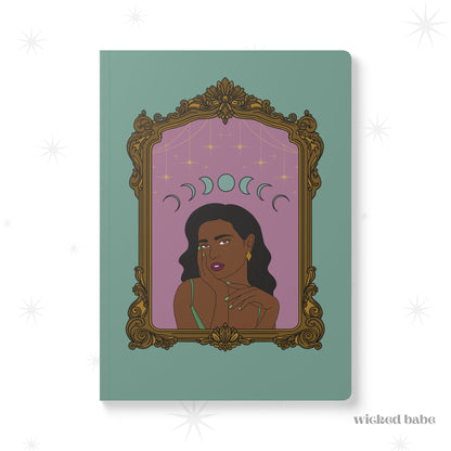 Front cover of green softcover journal notebook with design of a dark skinned woman with black hair wearing green on a pink background with moon phases above surrounded by ornate gold frame