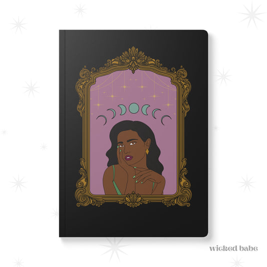 Front cover of black softcover journal notebook with design of a dark skinned woman with black hair wearing green on a pink background with moon phases above surrounded by ornate gold frame