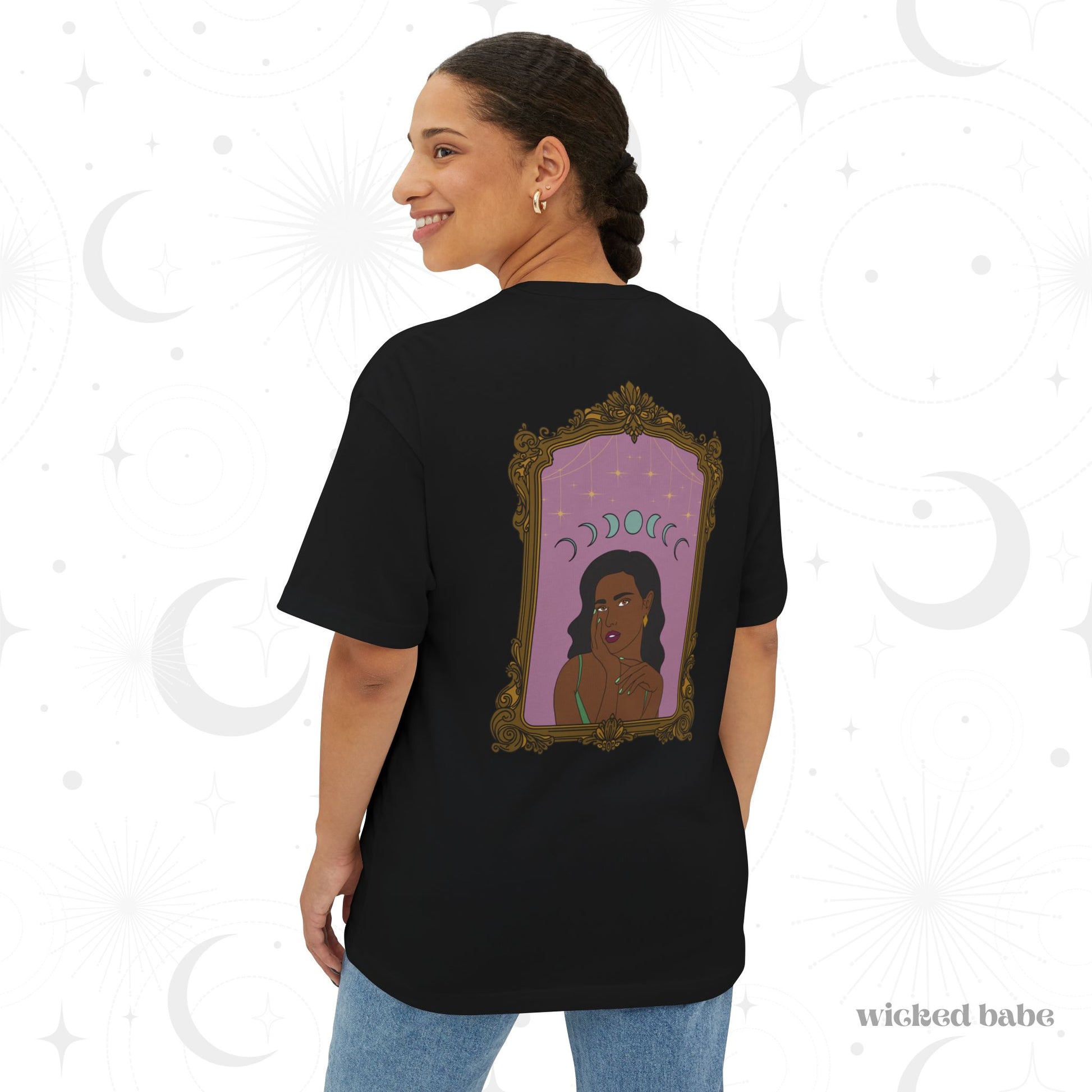 Woman wearing an oversized short sleeve black t-shirt with design of a dark skinned woman with black hair wearing green on a pink background with moon phases above surrounded by ornate gold frame on the back