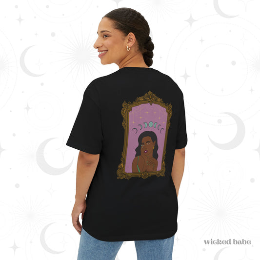 Woman wearing an oversized short sleeve black t-shirt with design of a dark skinned woman with black hair wearing green on a pink background with moon phases above surrounded by ornate gold frame on the back