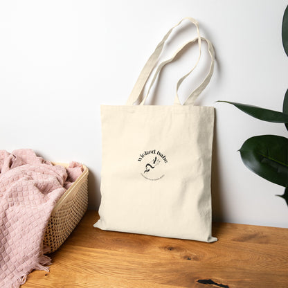The back of a canvas tote bag with black wicked babe hand with serpent logo in the center leaning against the wall