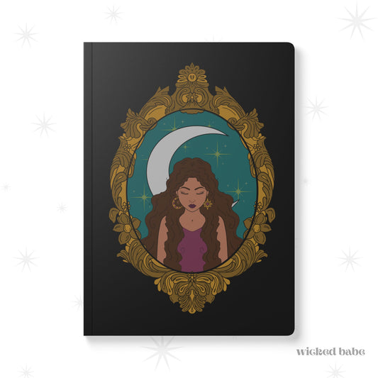 Front cover of black softcover journal notebook with design of a tan skinned woman with brown wavy hair wearing purple in front of large crescent moon on a teal background surrounded by ornate gold frame
