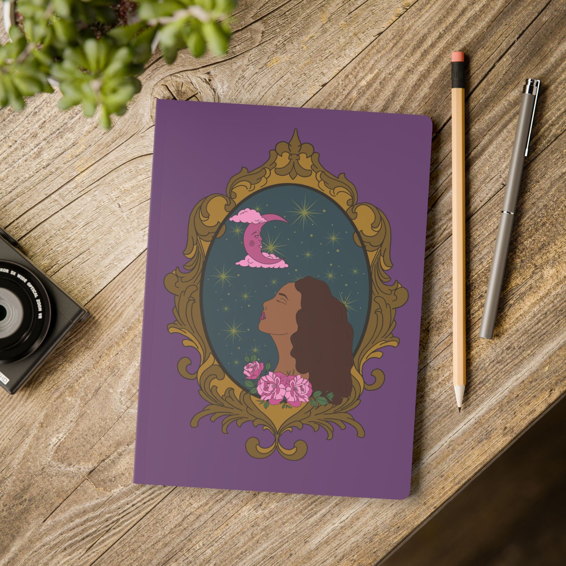 Front cover of purple softcover journal notebook with design of a tan skinned woman with red brown wavy hair with pink flowers and a pink crescent moon above on a teal background surrounded by an ornate gold frame on wooden table with pen and pencil