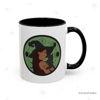 White mug with black accents, handle on the right, featuring a design of a woman wearing a witch's hat holding a gray cat with moon phases on a green background