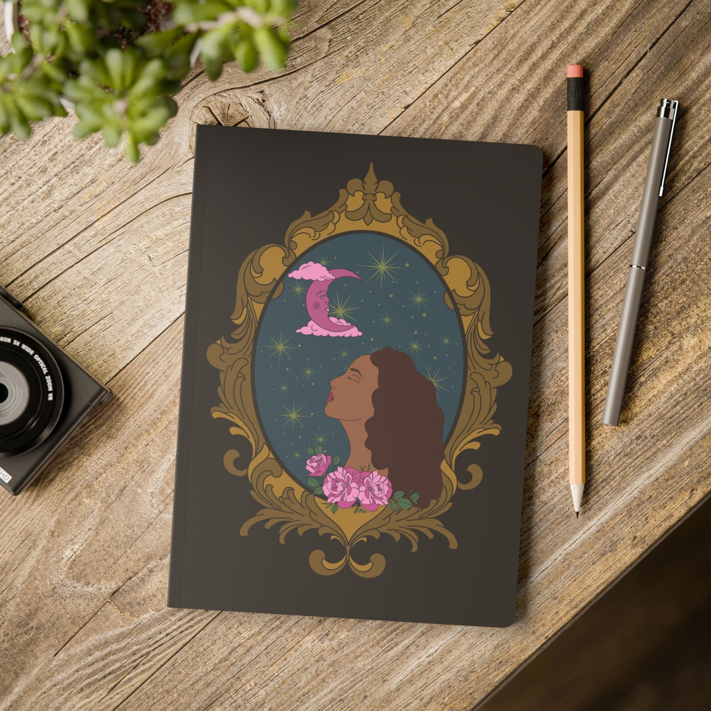 Front cover of black softcover journal notebook with design of a tan skinned woman with red brown wavy hair with pink flowers and a pink crescent moon above on a teal background surrounded by an ornate gold frame on wooden table with pen and pencil