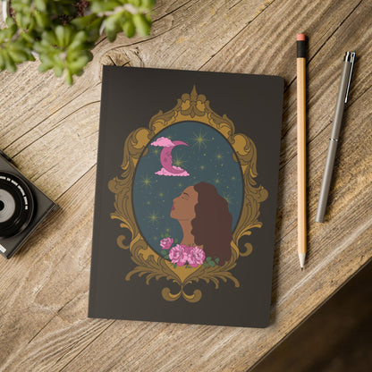 Front cover of black softcover journal notebook with design of a tan skinned woman with red brown wavy hair with pink flowers and a pink crescent moon above on a teal background surrounded by an ornate gold frame on wooden table with pen and pencil